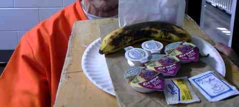 Individual in orange prison jumpsuit holds tray with overripe banana, three packages of grape jam, coffee and creamer packets, and opaque bag which may contain matzah