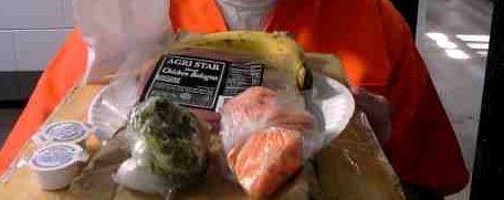 Individual in orange prison jumpsuit holds tray showing banana, package labeled "chicken bologna," two baggies with unidentifiable, uncooked foodstuffs  -- maybe vegetable and fruit.