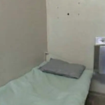 Prison cell with metal sink inches from cot covered with thin blanket and pillow