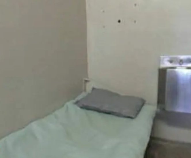 Prison cell with metal sink inches from cot covered with thin blanket and pillow